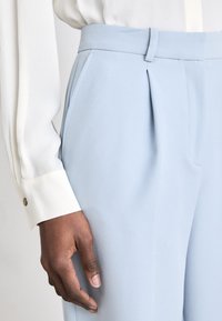 Pantalons sur mesure bleu clair avec plis sur les côtés et poches, en tissu lisse. La taille est structurée, accompagnée d'une blouse blanche à poignets.