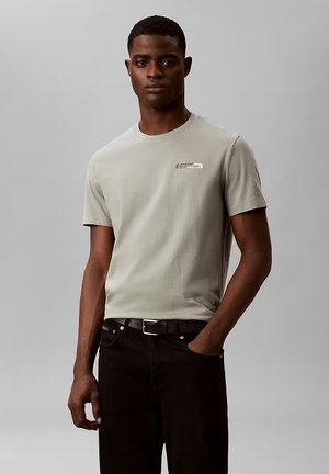 Uomo che indossa una t-shirt Calvin Klein grigio chiaro infilata in pantaloni neri, in piedi contro uno sfondo grigio uniforme con una mano in tasca.