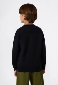 Bambino con capelli castani che indossa una maglietta nera a maniche lunghe e pantaloni verde oliva, girato di spalle su uno sfondo bianco uniforme.