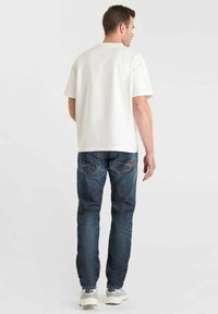 Uomo in piedi di spalle, che indossa una maglietta bianca a maniche corte, jeans blu e sneaker grigio chiaro su uno sfondo bianco.