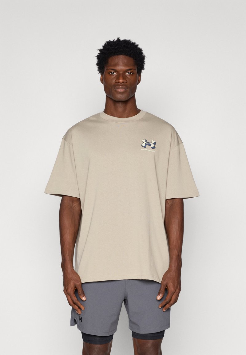 T-shirt in cotone beige con vestibilità rilassata, scollatura rotonda e un piccolo logo blu navy sul petto sinistro. Indossato con pantaloni corti grigi.