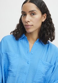 Camicia azzurra chiaro, con bottoni e colletto, due tasche frontali e un tessuto a trama. Presenta una vestibilità comoda e bottoni ben visibili.