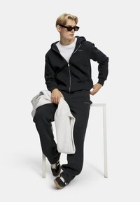 Marineblå hettegenser med glidelås og matchende bukser, med en hvit t-skjorte under. Strukturert stoff, uformell passform. Grå sweatshirt over leggen. Sorte sneakers.