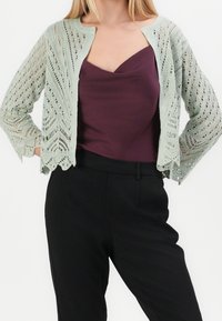 Häkelstrick-Cardigan in sanftem Grün, mit offenem Front, gewelltem Saum und kompliziertem Muster. Kombiniert mit einem tiefen lila Oberteil und schwarzen Hosen.