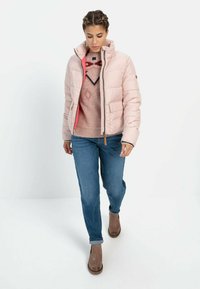 Lyserød pufferjakke med høj krave, lynlås og sidelommer, båret over en mønstret sweater og blå jeans med brune ankelstøvler.