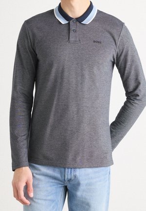 Homme portant un polo à manches longues gris foncé avec un col rayé bleu clair et blanc, et un jean bleu clair.