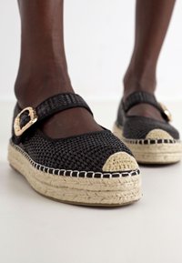 Čierne tkané espadrilky s okrúhlym otvoreným prstom, béžovou jute podrážkou a dekoratívnou zlatou sponou. Textúrovaný materiál s plochým dizajnom.