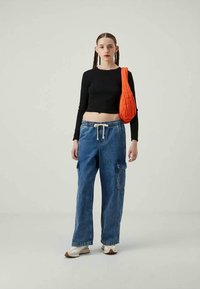 Young woman stands wearing black long-sleeve crop top, loose blue cargo jeans, white sneakers, and carries an orange quilted shoulder bag.