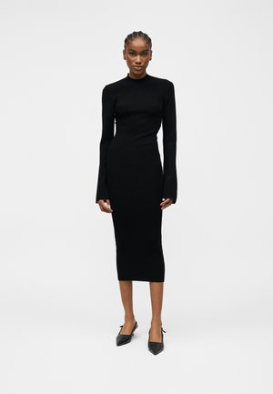 Kvinde med fletninger iført en tætsiddende sort midikjole med lange ærmer og sorte spidse slingback-sko, stående mod en ensfarvet baggrund.