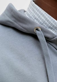 Sweat à capuche gris clair en tissu texturé, doté d'un cordon de serrage et d'œillets métalliques. En dessous, on aperçoit une chemise à col bleu et blanc à carreaux.