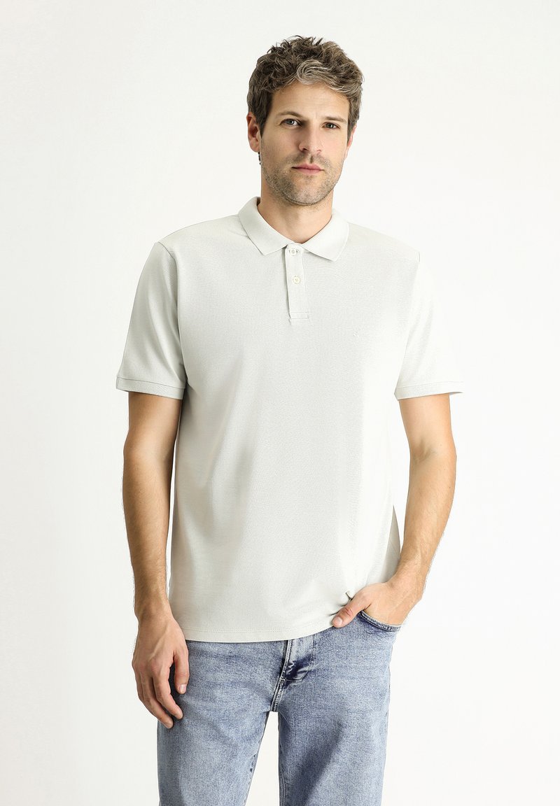 Polo shirt grigio chiaro con colletto e chiusura a tre bottoni. Maniche corte, tessuto testurizzato e vestibilità rilassata abbinata a jeans blu.