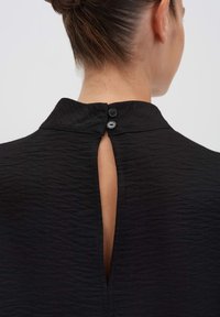 Schiena di una donna con i capelli raccolti, che indossa un top nero strutturato con un alto colletto e due bottoni neri che fissano una fessura verticale.