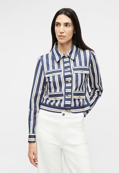 Γυναίκα με μακριά σκούρα μαλλιά φοράει μπλε και άσπρο κάθετο ριγέ σακάκι με χρυσά κουμπιά και άσπρο παντελόνι, στέκεται μπροστά σε απλό φόντο.