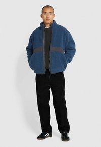 Giacca in pile blu con una banda orizzontale grigia. Presenta un collo alto, chiusura con zip e due tasche frontali. Abbinata a pantaloni neri.