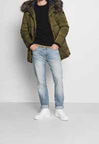 Vojenská zelená zateplená parka s kapucí s kožešinovým lemem, černý svetr, světlé modré džíny a bílé tenisky. Neformální outfit s kontrastními texturami.