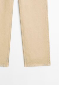 Beżowe dżinsy o prostych nogawkach, z przeszytymi szwami i lekko postrzępionym dołem, na białym tle.