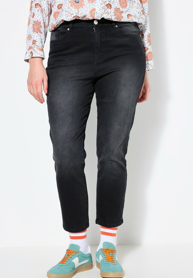 Jeans de mezclilla negra con corte ajustado, que cuentan con cinco bolsillos, cierre de botón y cremallera, y una textura desgastada, combinados con zapatillas coloridas.
