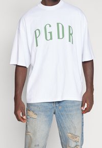 Vit oversize T-shirt med grön text "PGDR". Har rundad halsringning och korta ärmar. Parat med slitna ljusblå jeans.