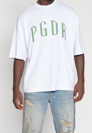Bílá oversized tričko s zeleným textem "PGDR". Má kulatý výstřih a krátké rukávy. Spárováno s obnošenými světle modrými džíny.