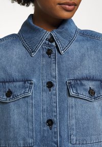 Camisa de mezclilla en azul claro con un cuello estructurado, botones negros y dos bolsillos en el pecho. Presenta una textura suave y detalles de costura visibles.