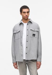 Camisa de franela gris con un diseño de botones, dos bolsillos frontales y un parche de marca negro. Llevada sobre una camiseta blanca, combinada con pantalones gris claro.