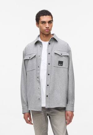 Grijs flanellen shirt met een knoopsluiting, twee voorkruiszakken en een zwart merklabel. Draag het over een wit T-shirt, gecombineerd met lichtgrijze broek.