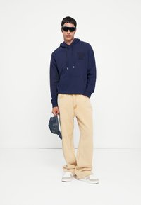 Navy blue hoodie with logo detail, beige wide-leg pants, gray cap in hand, white sneakers, and sunglasses. Model stands against plain background.