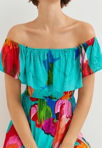 Robe décolletée en turquoise vibrant avec des motifs floraux rouge, rose et vert ; tissu léger avec un col froncé.