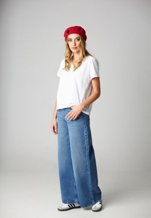 Kobieta w czerwonym berecie, białej koszulce, szerokich niebieskich dżinsach i białych trampkach, stojąca na tle jednolitego szarego tła.