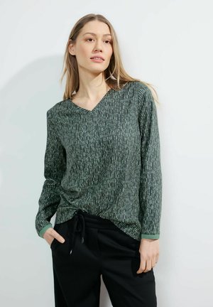 Haut à manches longues vert avec un col en V, présentant un motif vertical d'accents vert clair sur un tissu texturé. Associé à un pantalon noir.