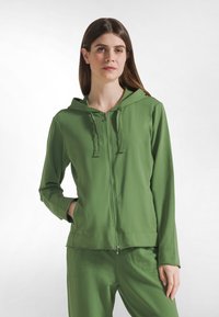 Sudadera verde con capucha, cierre de cremallera, mangas largas y bolsillos delanteros. Hecha de un material suave y texturizado, con un dobladillo recto y detalles con cremallera.