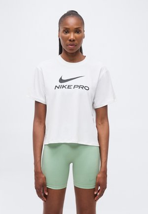 Kobieta z związanymi włosami, ubrana w białą koszulkę Nike Pro i jasnóżółte dopasowane krótkie spodenki sportowe, stojąca na jednolitym tle.