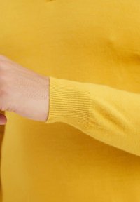 Bras portant un pull jaune à manches longues avec un poignet côtelé, poing partiellement serré visible sur un fond jaune uni.