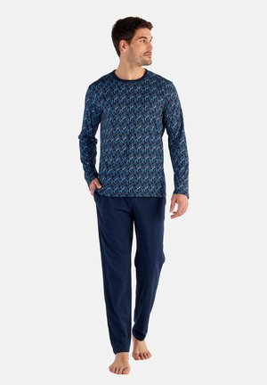 Man die een blauw patroon lange mouwen shirt en effen marineblauwe broek draagt, blootsvoets loopt tegen een eenvoudige lichte achtergrond.