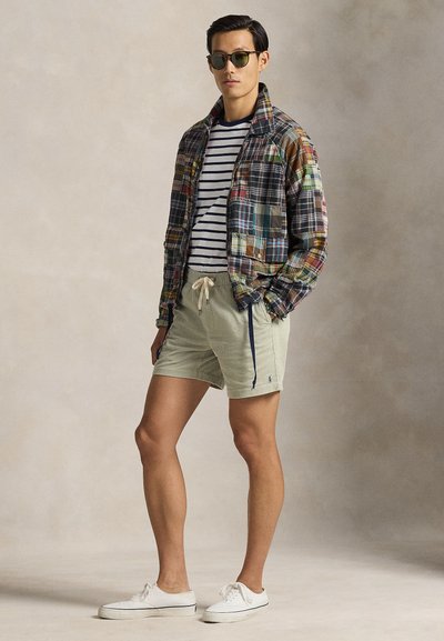 Πολύχρωμο καρό μπουφάν με δύο τσέπες, πουκάμισο με ρίγες, ανοιχτό πράσινο σορτς και λευκά αθλητικά παπούτσια, ολοκληρωμένο με στρογγυλά γυαλιά ηλίου.