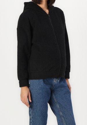 Kobieta w czarnej polarowej kurtce z zamkiem i kapturem oraz niebieskich dżinsach, stojąca na jednolitym białym tle, kadrowana na wysokości ramion i ud.