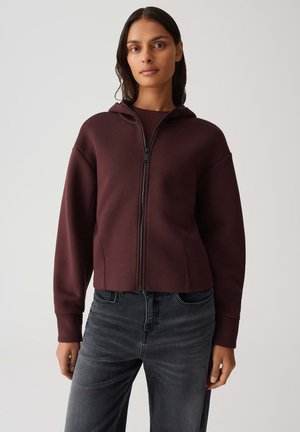 Burgunderfarbene Zip-Hoodie mit taillierter Passform, großer Kapuze und gerippten Bündchen. Kombiniert mit dunkelgrauen, weiten Jeans. Glatte Textur.