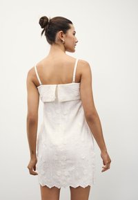 Robe blanche brodée de motifs floraux, avec des bretelles fines réglables et un ourlet festonné. Le matériau semble doux avec une finition texturée.