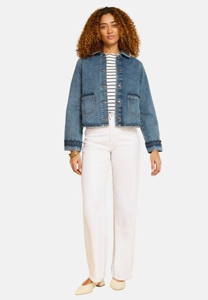 Vrouw met krullend haar die een blauwe spijkerjas draagt over een gestreept shirt, witte high-waisted broek, beige schoenen en gouden armbanden, staand op een witte achtergrond.