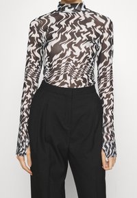 Donna che indossa un top lungo a maniche trasparenti con un motivo astratto bianco e nero, infilato in pantaloni neri a gamba larga con vita alta.