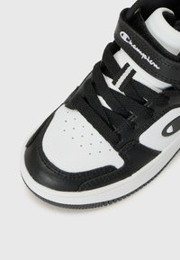 Baskets en noir et blanc avec une tige texturée, un laçage prononcé et un logo sur la sangle, centrées sur la zone des orteils.