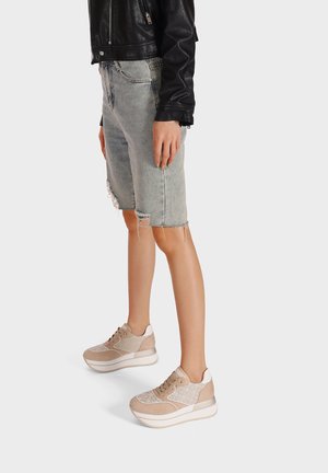 Sneakers platform beige con tessuto texture, dettagli in rete e suole bianche. Indossati con pantaloncini di jeans strappati e una giacca di pelle nera.