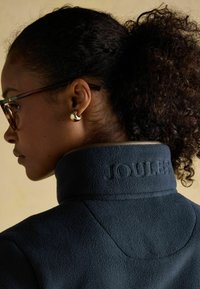 Žena s kučeravými vlasmi stiahnutými dozadu, nosí okrúhle hrubé okuliare z korytnačiny, zlaté kruhové náušnice a námornícky fleecový kabát s vyšitým nápisom "JOULES" na golieri.