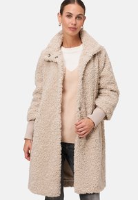 Beige nepbontjas met een gelapt kraag, knoopsluiting aan de voorkant en brede mouwen, gestapeld over een licht V-hals trui.