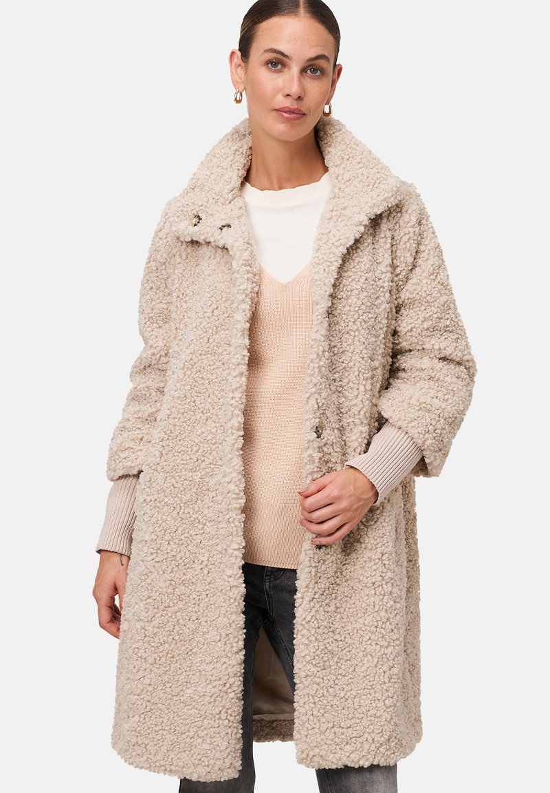Beige nepbontjas met een gelapt kraag, knoopsluiting aan de voorkant en brede mouwen, gestapeld over een licht V-hals trui.