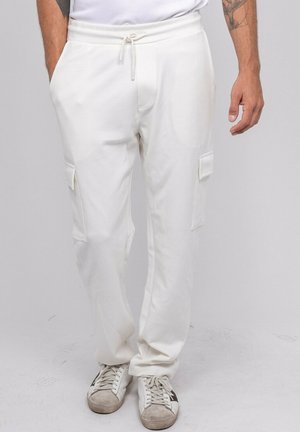 Homme debout, les mains dans les poches, vêtu d’un pantalon cargo jogger blanc, de baskets blanches et d’une chemise blanche ; tatouage visible sur l’avant-bras droit.