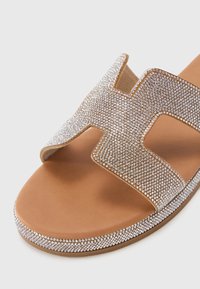 Sandal med gyldne stropper, prydet med klare rhinestones, og en tan fotseng i lær, med en flat, jevn såle.
