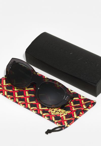 Black oversized cat-eye sunglasses on a patterned red and yellow pouch, with a textured black case featuring embossed brand name.