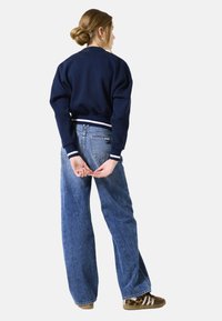 Sweatshirt bleu marine avec des poignets rayés blancs, associé à un jean ample bleu clair et des baskets marron avec des rayures blanches.