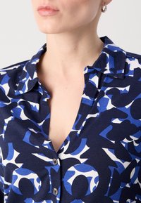 Femme portant une chemise à motif abstrait bleu et blanc avec col, partiellement déboutonnée, et une petite boucle d'oreille dans l'oreille gauche.